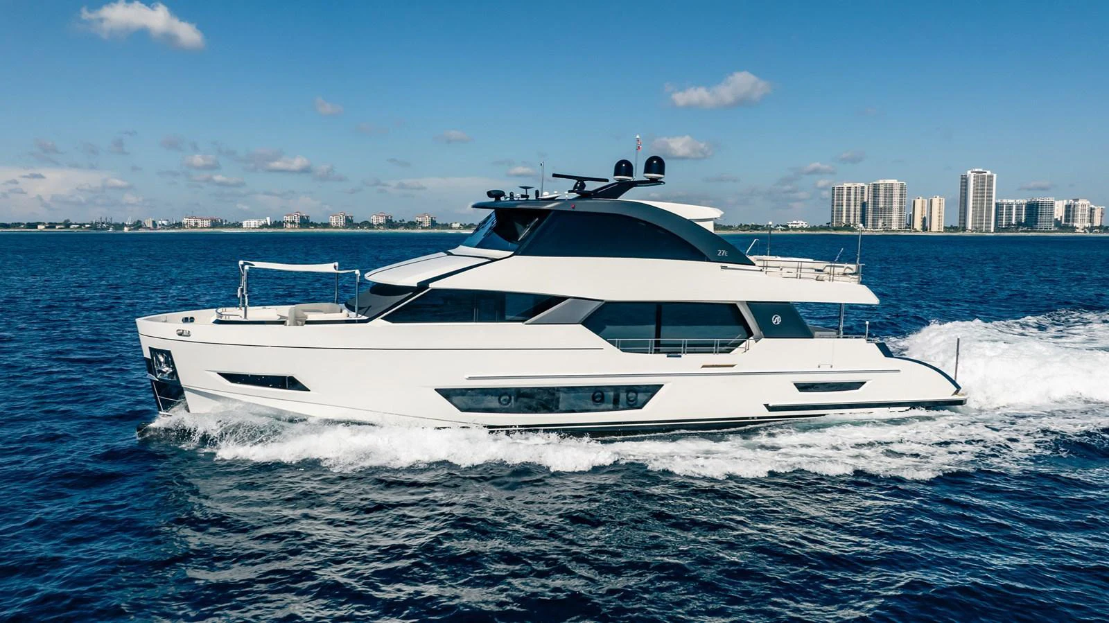 A sleek and modern yacht cruises through calm blue waters with a cityscape in the background. aboard AQUASITION Yacht for Sale