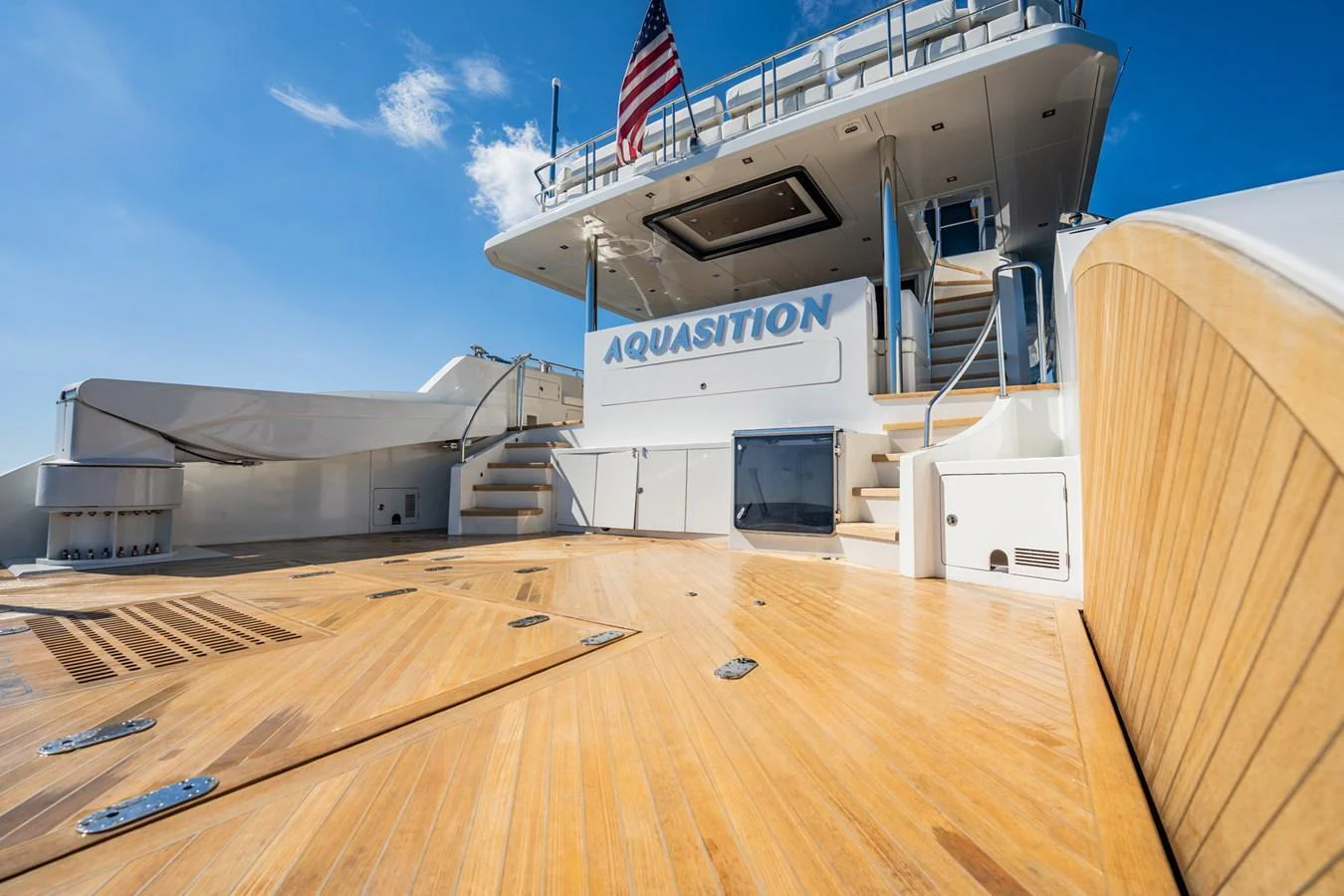 The aft deck features a spacious teak platform with steps leading up to the main deck, complemented by a sleek design and an American flag flying above. aboard AQUASITION Yacht for Sale