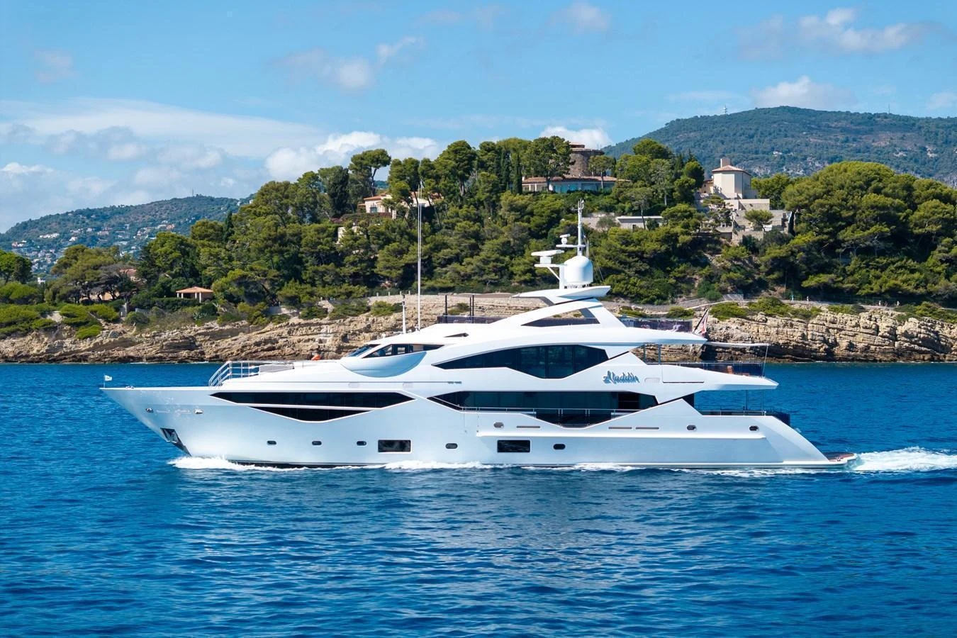 A sleek, modern yacht cruises through clear blue waters with a scenic coastal backdrop. aboard ALADDIN Yacht for Sale