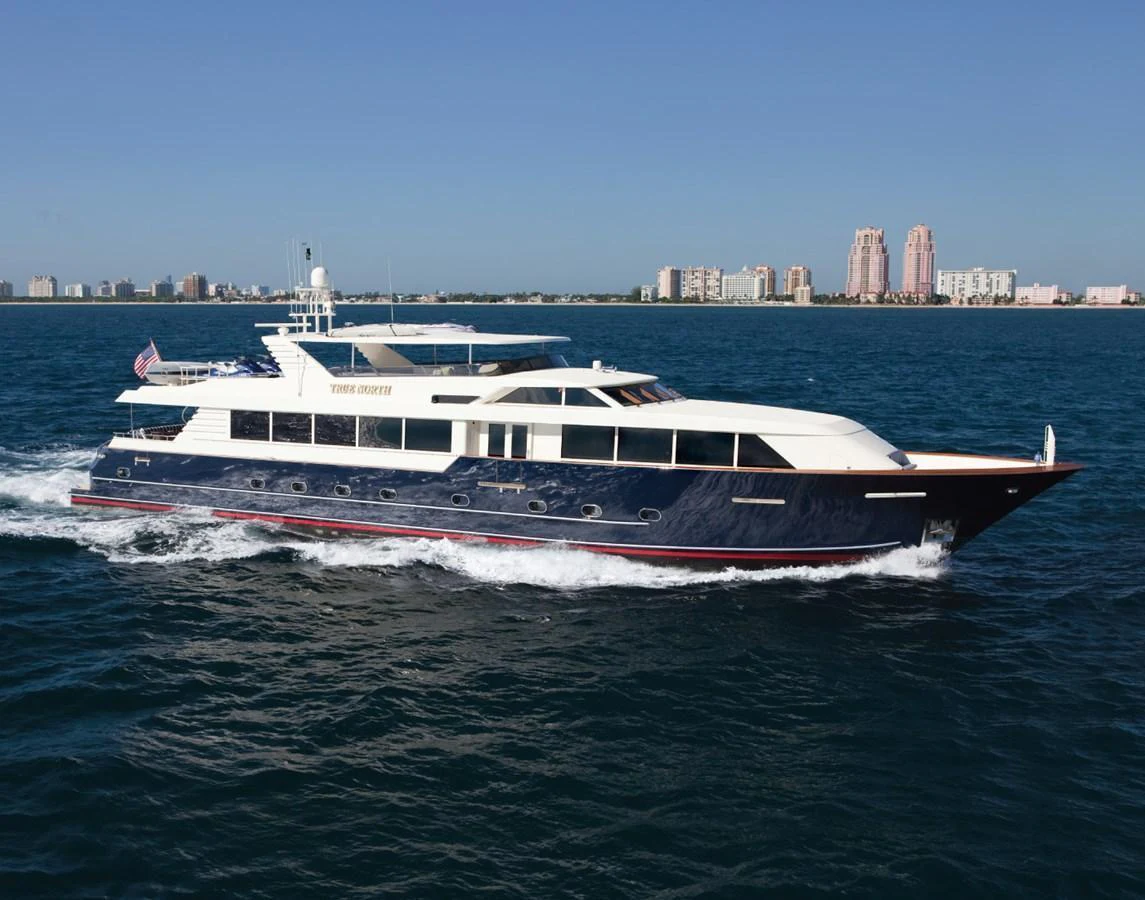 A sleek yacht cruises through the water with a cityscape in the background. aboard TRUE NORTH Yacht for Sale