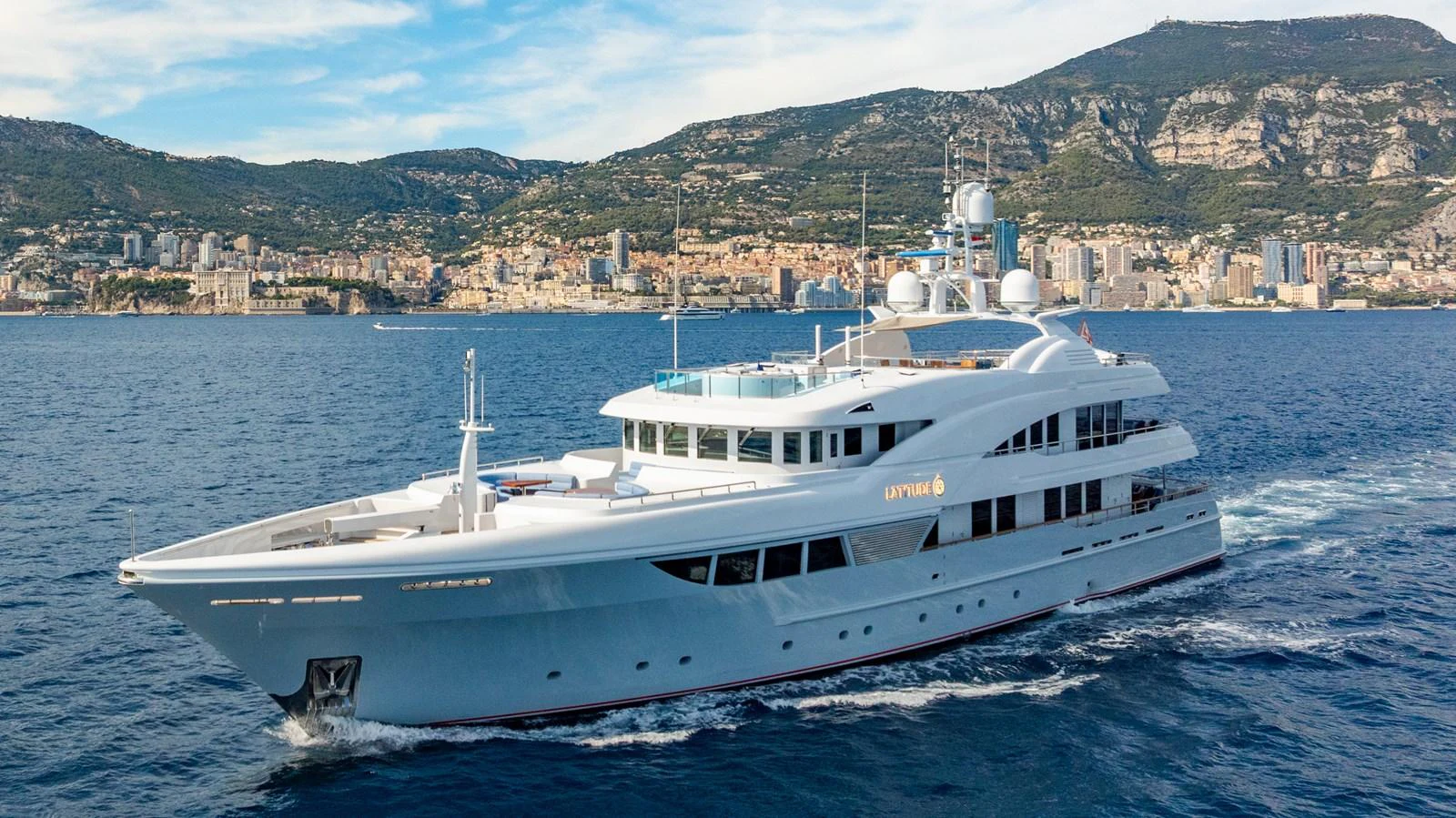 A luxurious yacht cruises along the coastline with a scenic city and mountainous backdrop. aboard LATITUDE Yacht for Sale