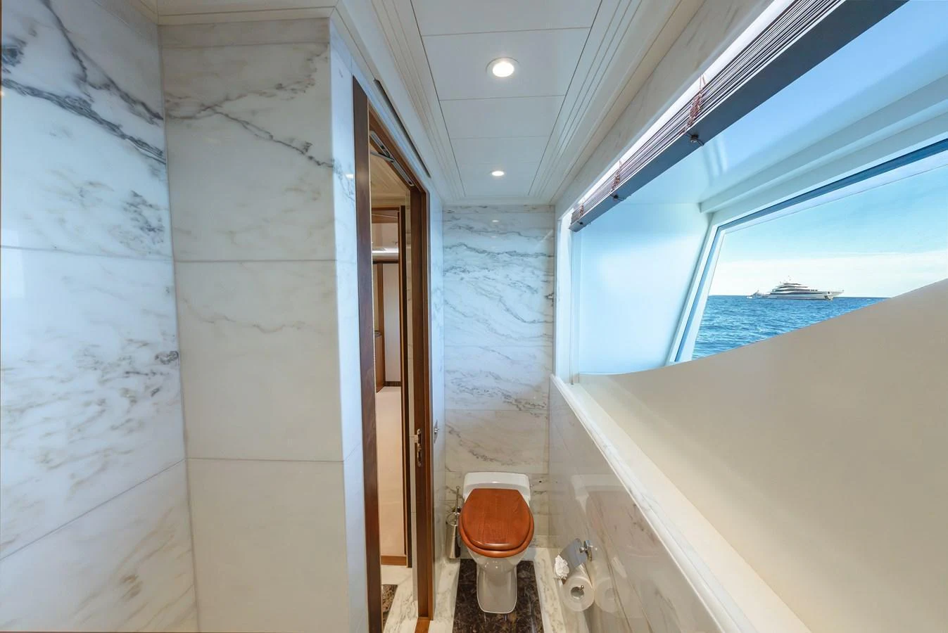 A luxurious bathroom with marble walls and a wooden toilet seat, featuring a window with an ocean view. aboard LATITUDE Yacht for Sale