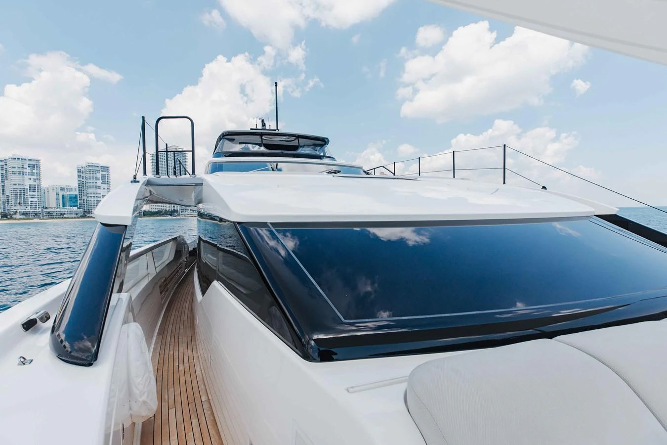 A sleek and modern yacht exterior with expansive windows and a teak deck, set against a backdrop of a city skyline and blue skies. aboard CHAMELEON Yacht for Sale