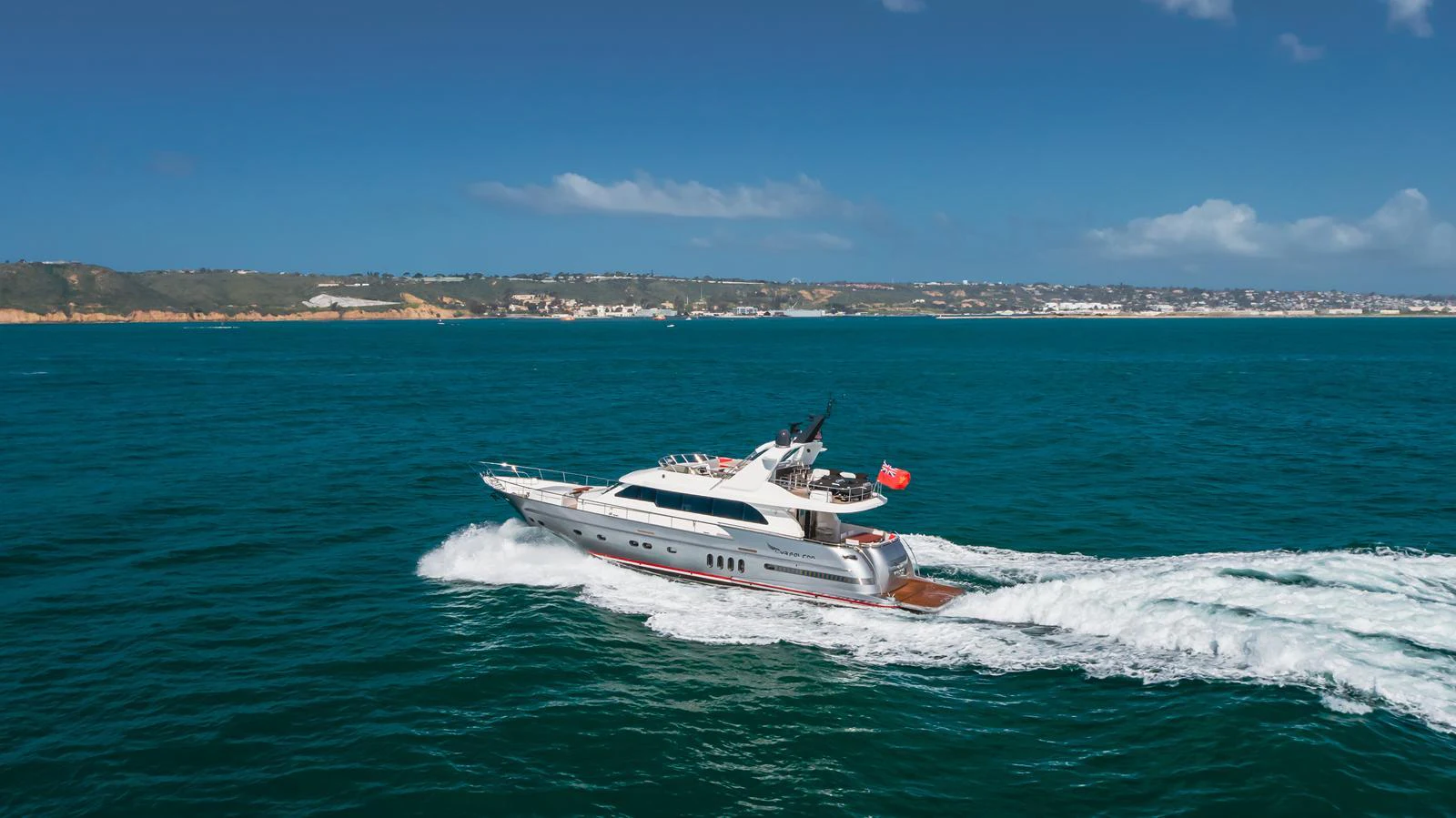 A sleek yacht cruises through vibrant blue waters with a scenic coastline in the background. aboard TEST NAME Yacht for Sale