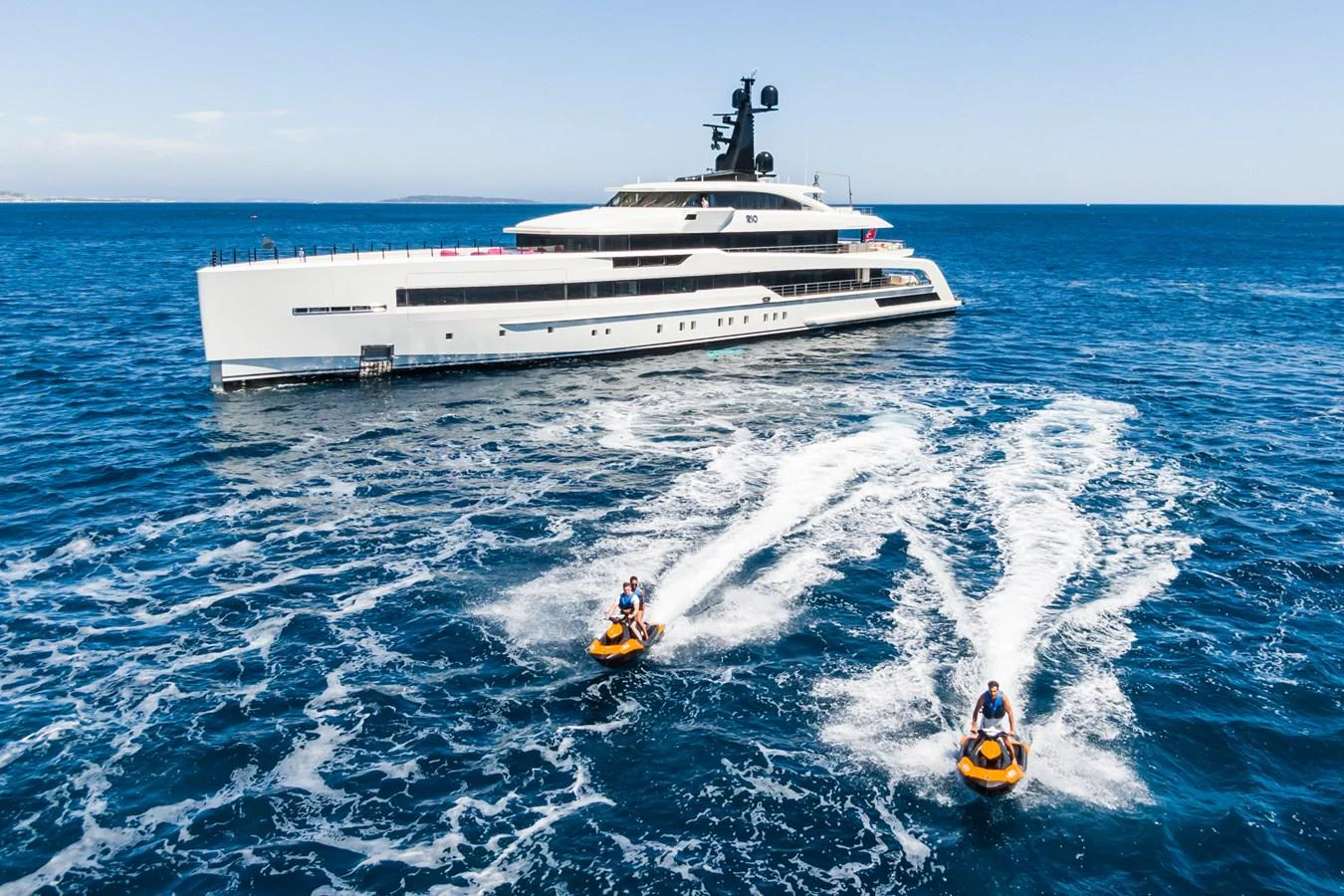 A sleek, modern yacht glides through the open sea with two jet skis trailing behind. aboard RIO Yacht for Sale