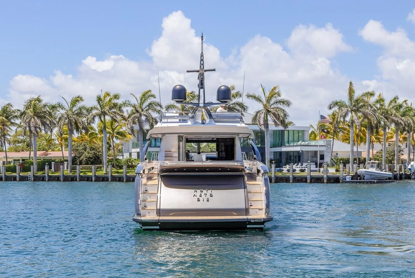 The yacht is seen from the rear, docked in a serene waterfront setting with palm trees and a clear sky in the background. aboard NO TIME TO DIE Yacht for Sale