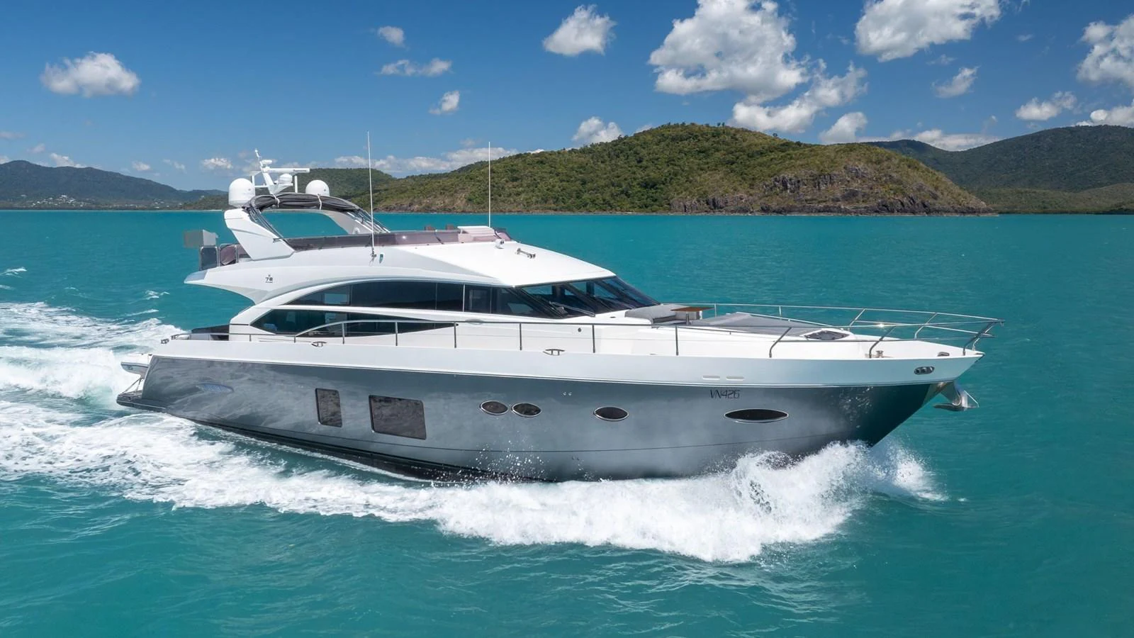 A sleek and modern yacht cruising through turquoise waters against a backdrop of lush hills. aboard ABOUT TIME Yacht for Sale