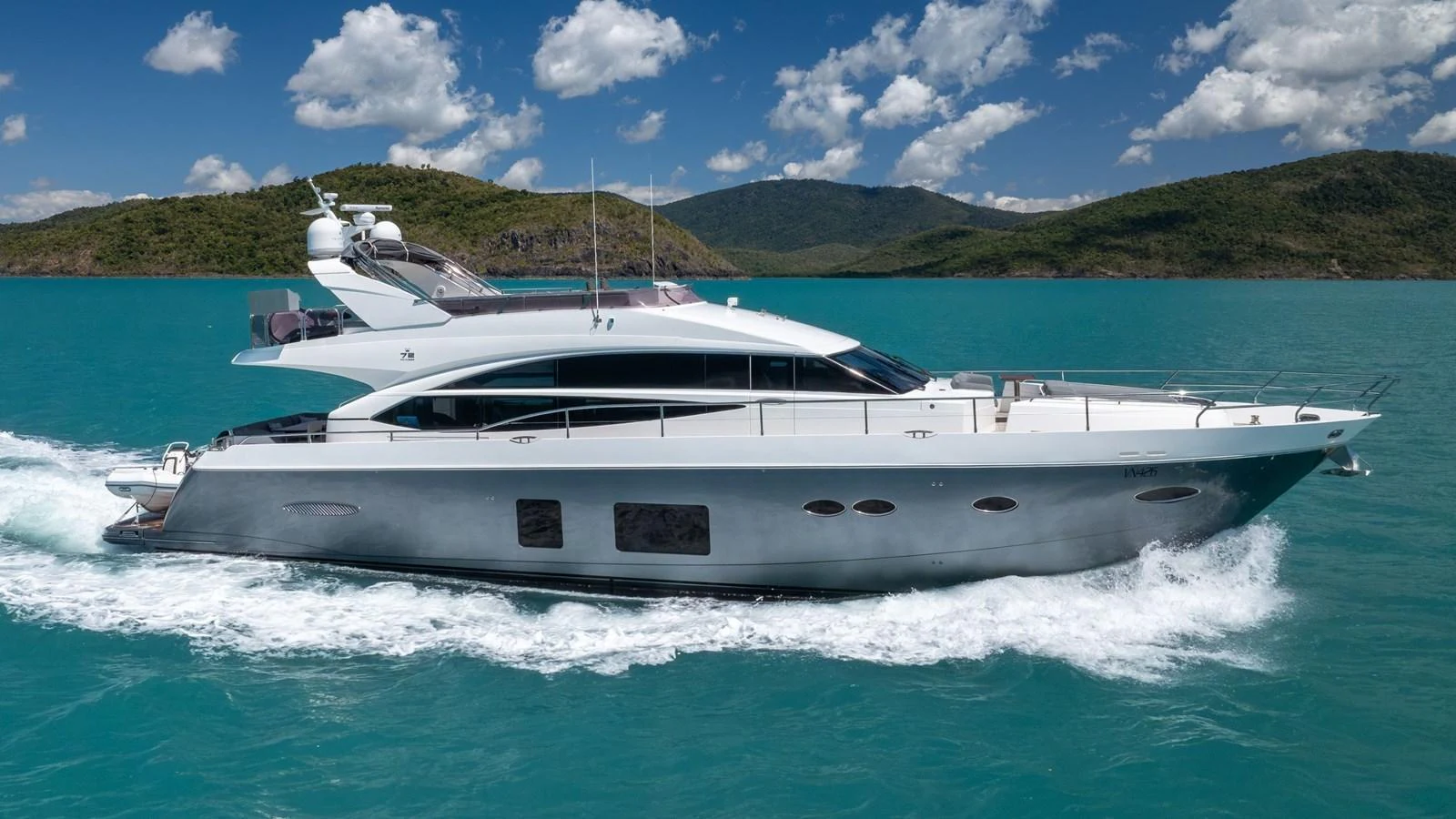 A sleek and modern yacht cruises through turquoise waters against a backdrop of lush hills and a bright blue sky. aboard ABOUT TIME Yacht for Sale