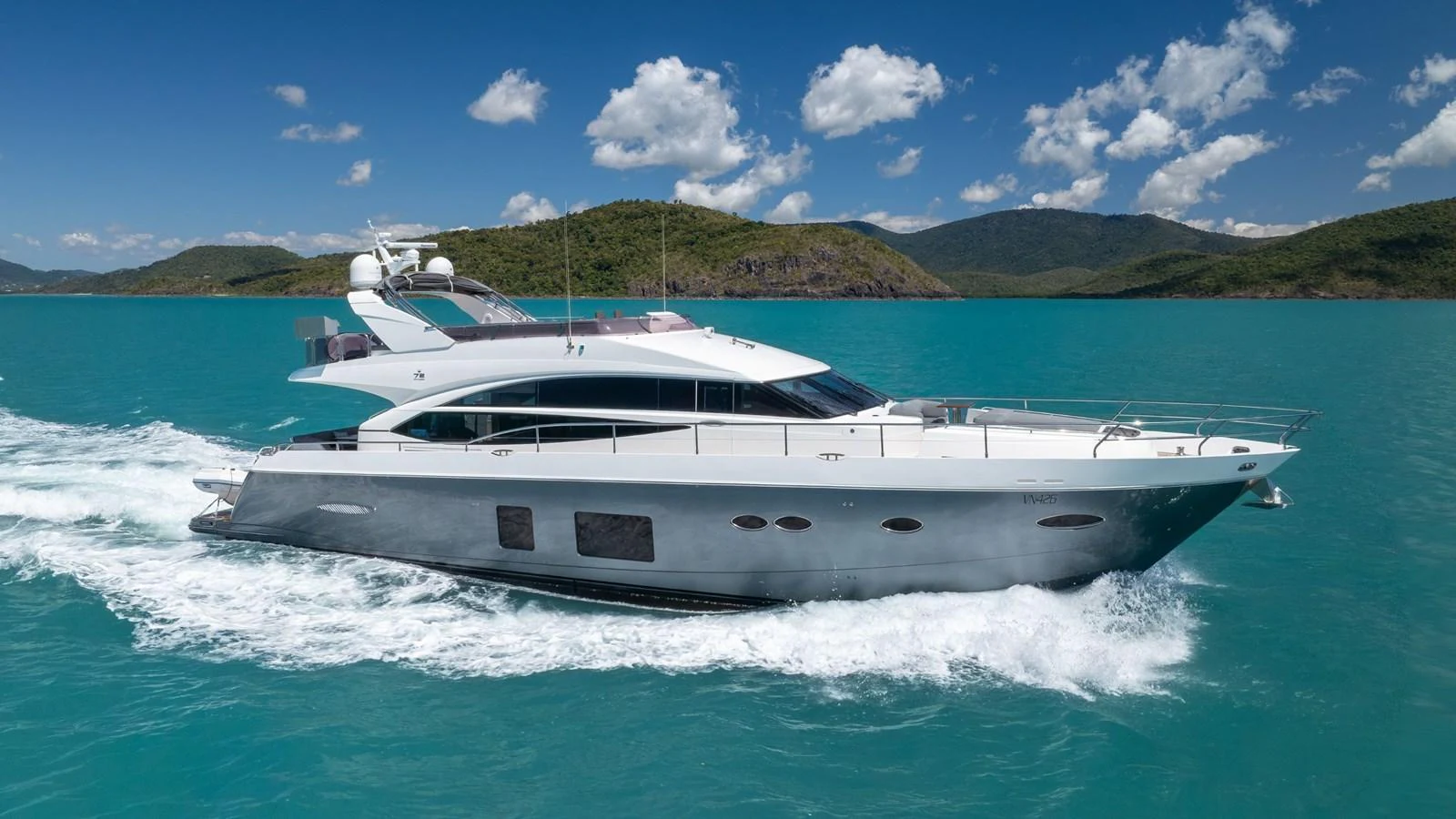 A sleek and modern yacht cruises through turquoise waters against a backdrop of lush green hills. aboard ABOUT TIME Yacht for Sale