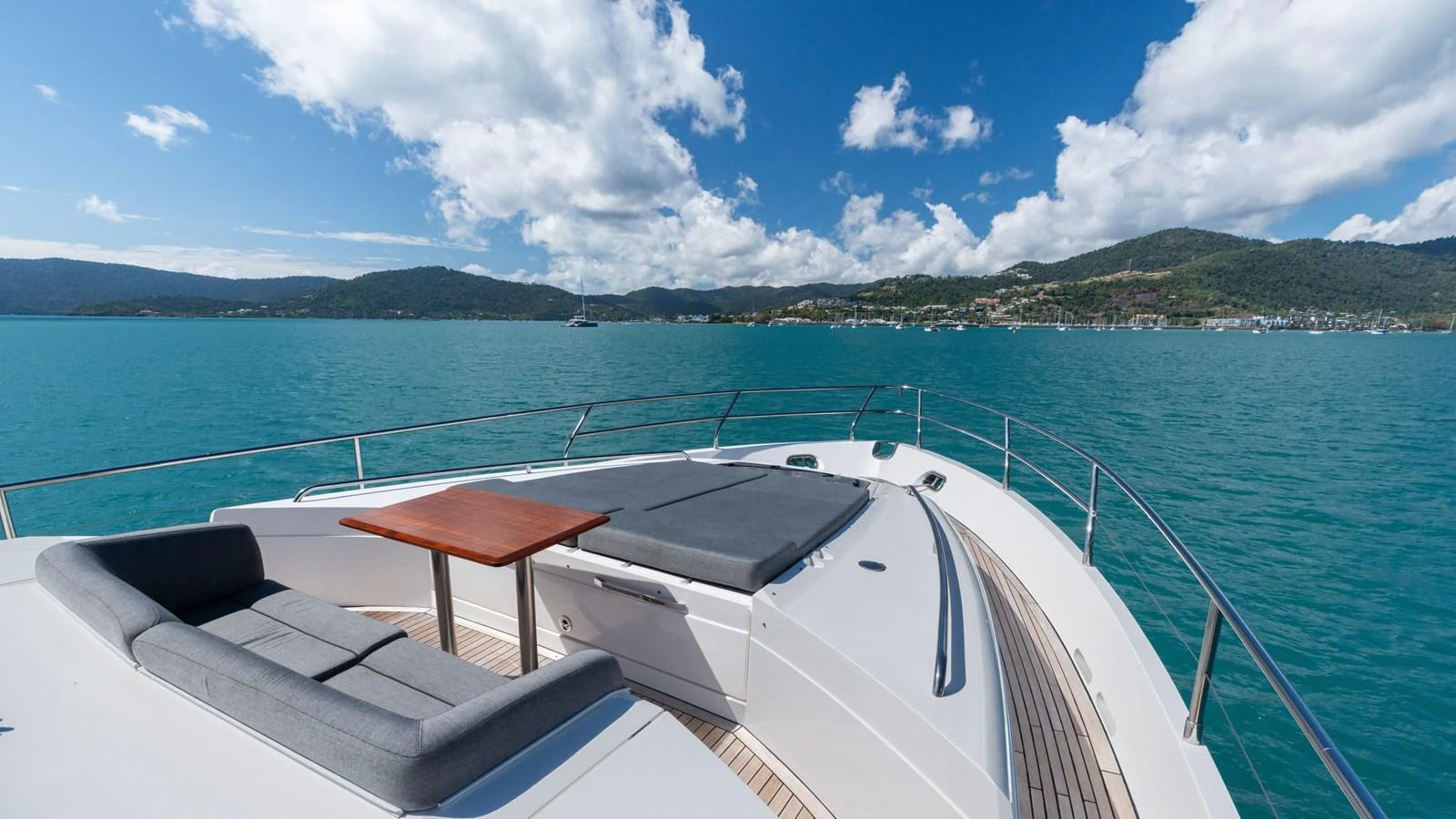 The yacht's foredeck features a spacious sunbathing area with plush cushions and a small table, set against a stunning seascape. aboard ABOUT TIME Yacht for Sale