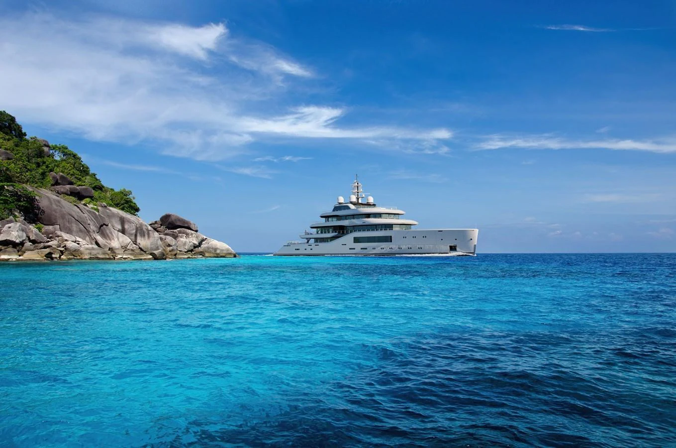 A luxurious yacht cruises through pristine turquoise waters near a rocky shoreline under a clear blue sky. aboard AMELS 80 Yacht for Sale