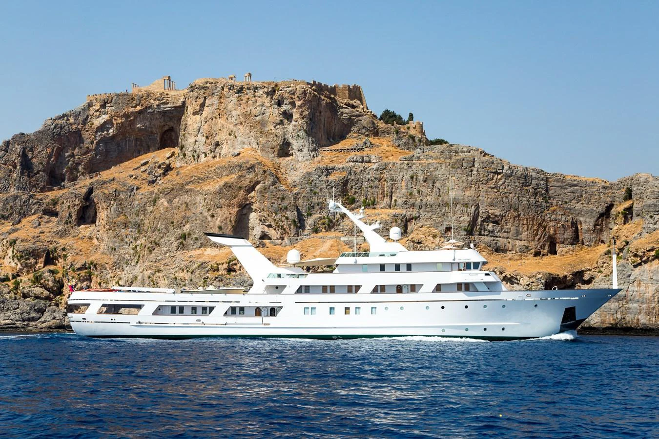 A sleek white yacht cruises along a rocky coastline under a clear blue sky. aboard ESMERALDA Yacht for Sale
