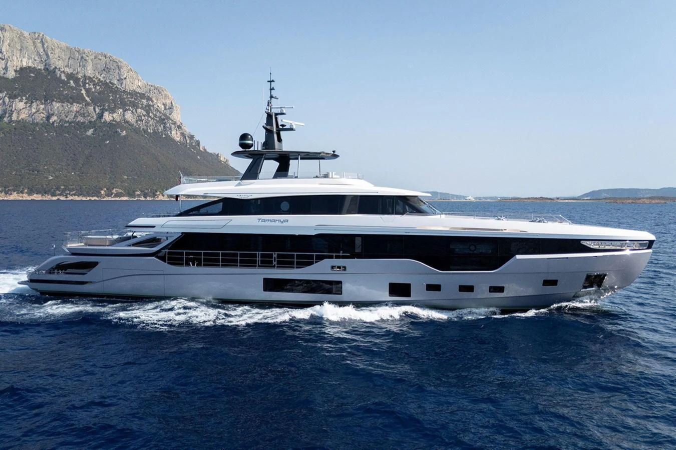 A sleek and modern yacht cruising on the open sea with a scenic mountainous backdrop. aboard TAMANYA Yacht for Sale