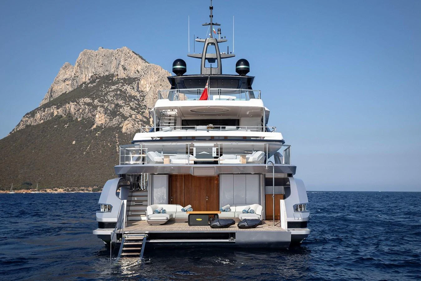 A luxurious yacht anchored near a scenic rocky coastline, showcasing its spacious aft deck with lounging areas and steps leading to the water. aboard TAMANYA Yacht for Sale