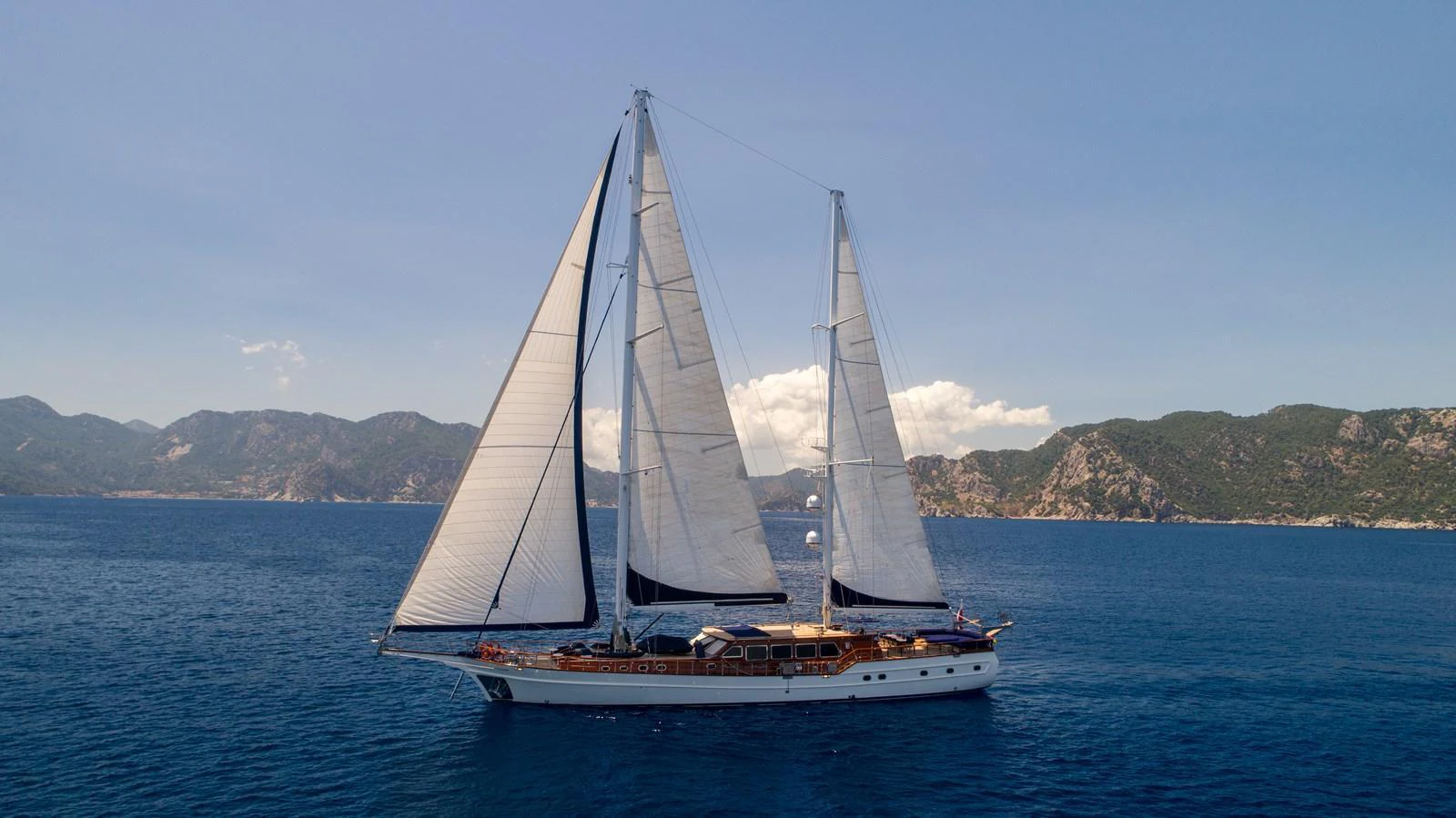 A stunning sailing yacht glides gracefully on the open sea with majestic mountains in the background. aboard VOYAGE Yacht for Sale