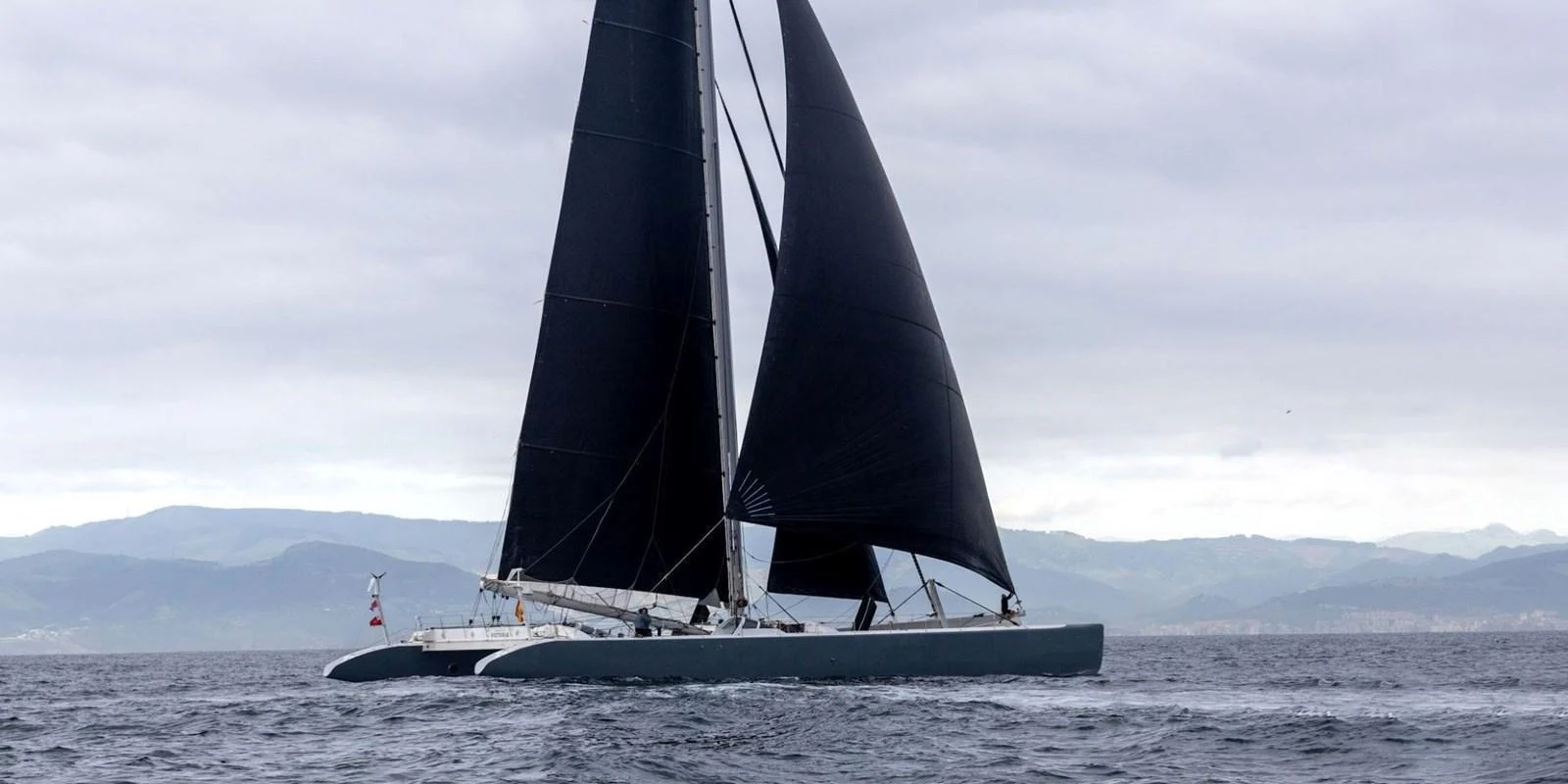 A sleek sailing yacht with dark sails glides through the water under a cloudy sky. aboard MAXICAT VICTORIA Yacht for Sale