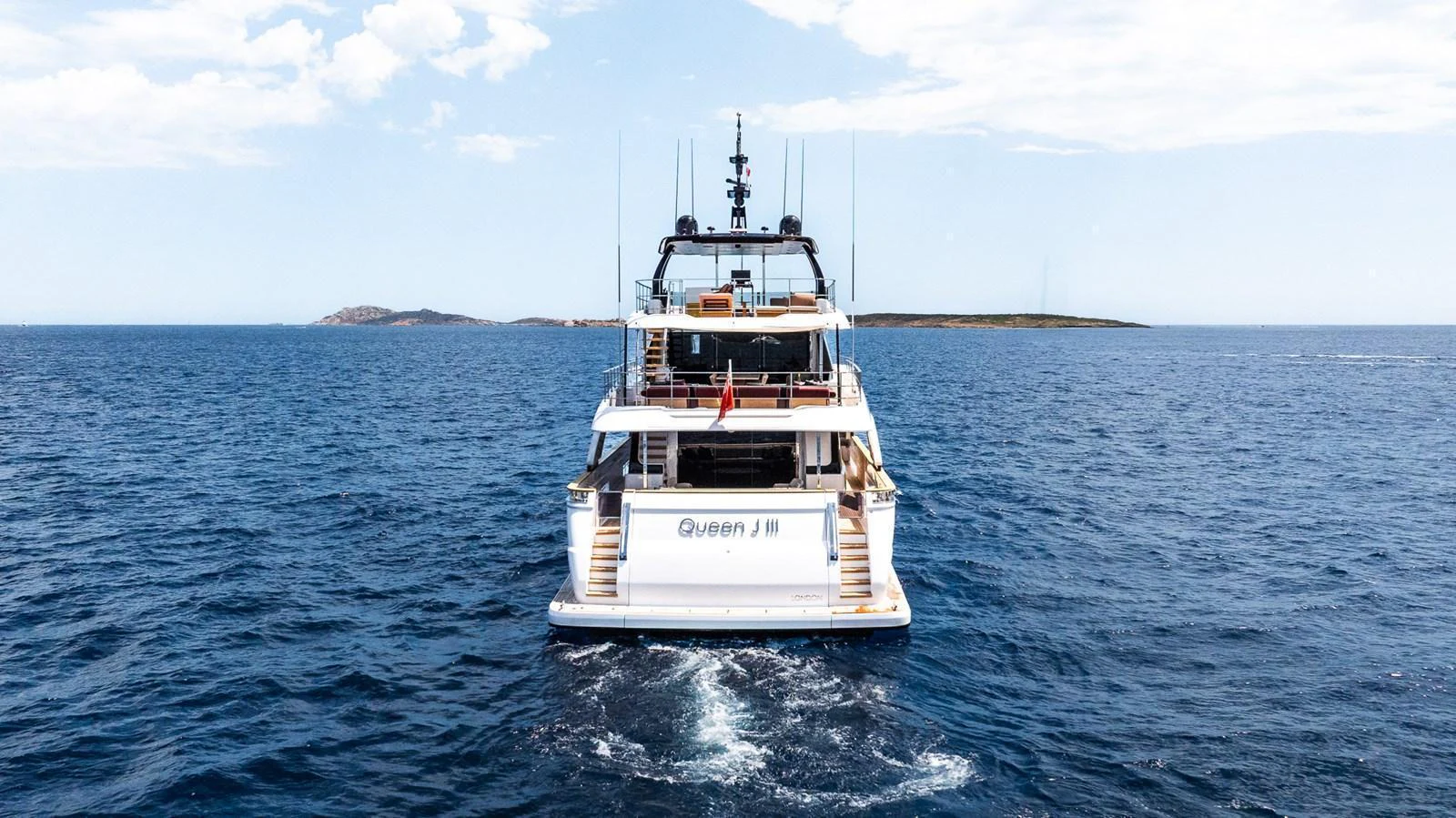 The yacht is seen from the rear, cruising through calm blue waters with a clear sky above. aboard QUEEN J III Yacht for Sale