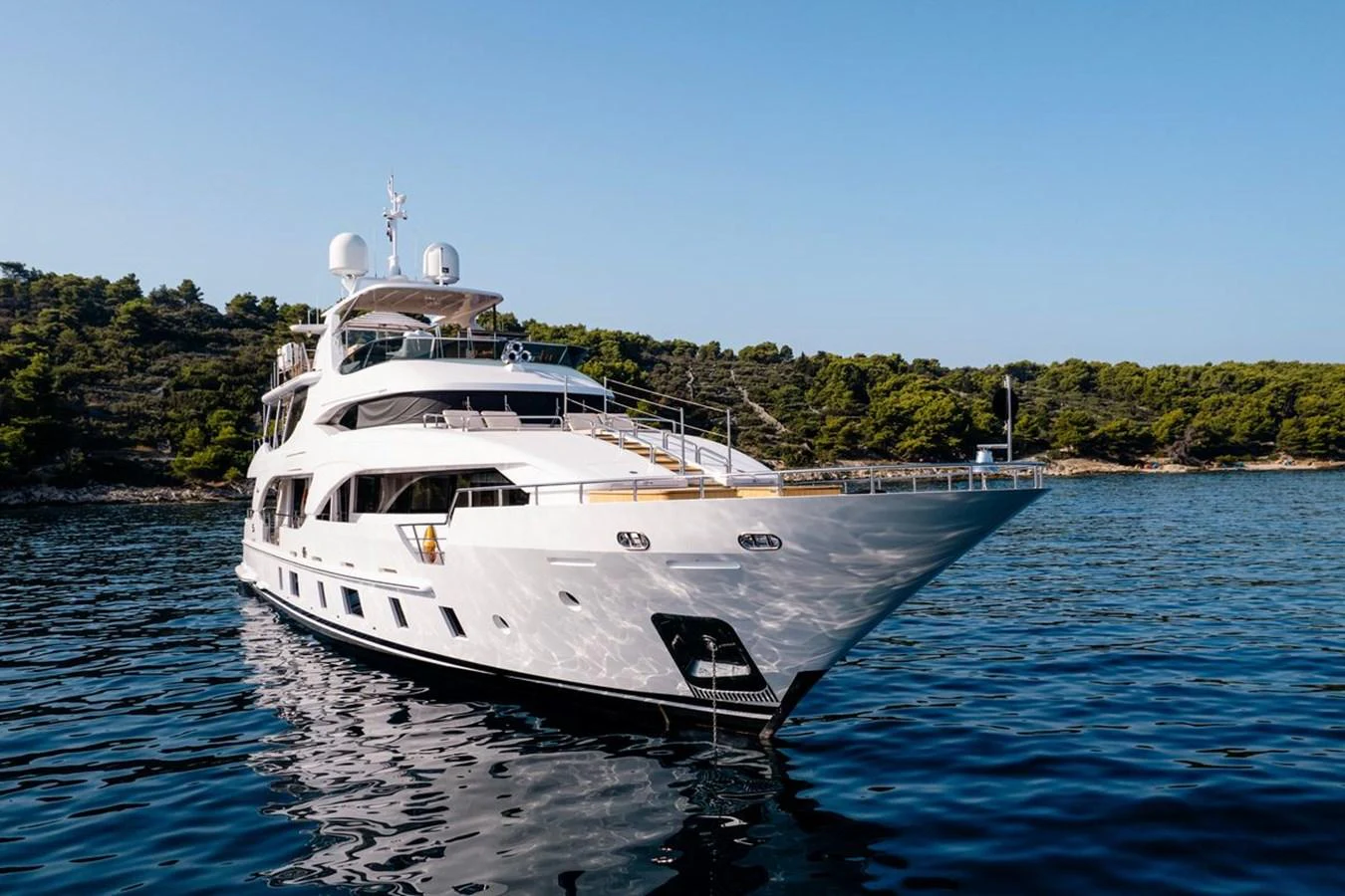 A luxurious yacht is anchored in calm, clear waters with a scenic forested coastline in the background. aboard AFINA Yacht for Sale