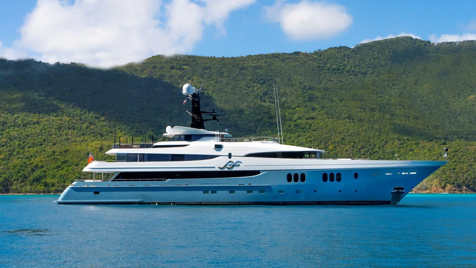 A sleek and luxurious yacht is anchored against a backdrop of lush green hills and clear blue waters. aboard SCOTT FREE Yacht for Sale