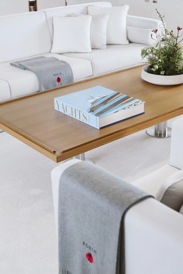 The salon features a cozy seating area with plush white sofas and a stylish wooden coffee table adorned with a decorative book and a floral arrangement. aboard RONIN Yacht for Sale