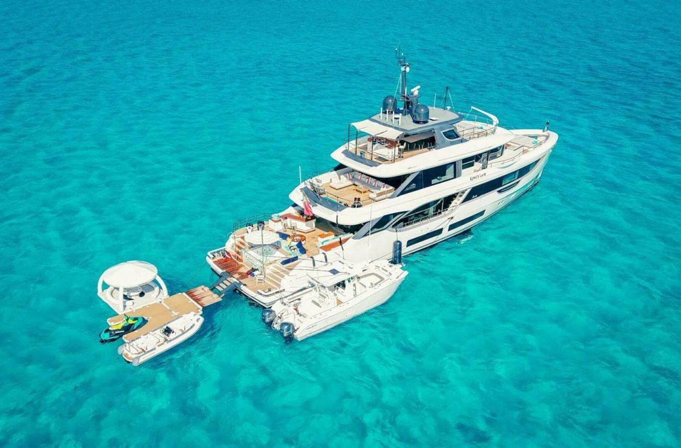 A luxurious yacht anchored in crystal-clear turquoise waters, accompanied by smaller boats and water toys. aboard KING'S LAIR Yacht for Sale