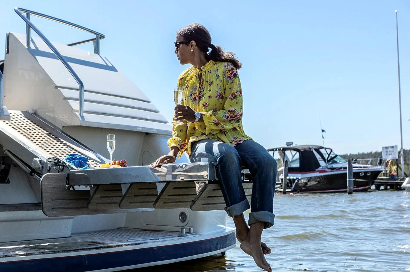 A person relaxes with a glass of champagne on the swim platform of a yacht, enjoying a sunny day by the water. aboard UNITED SPIRIT Yacht for Sale