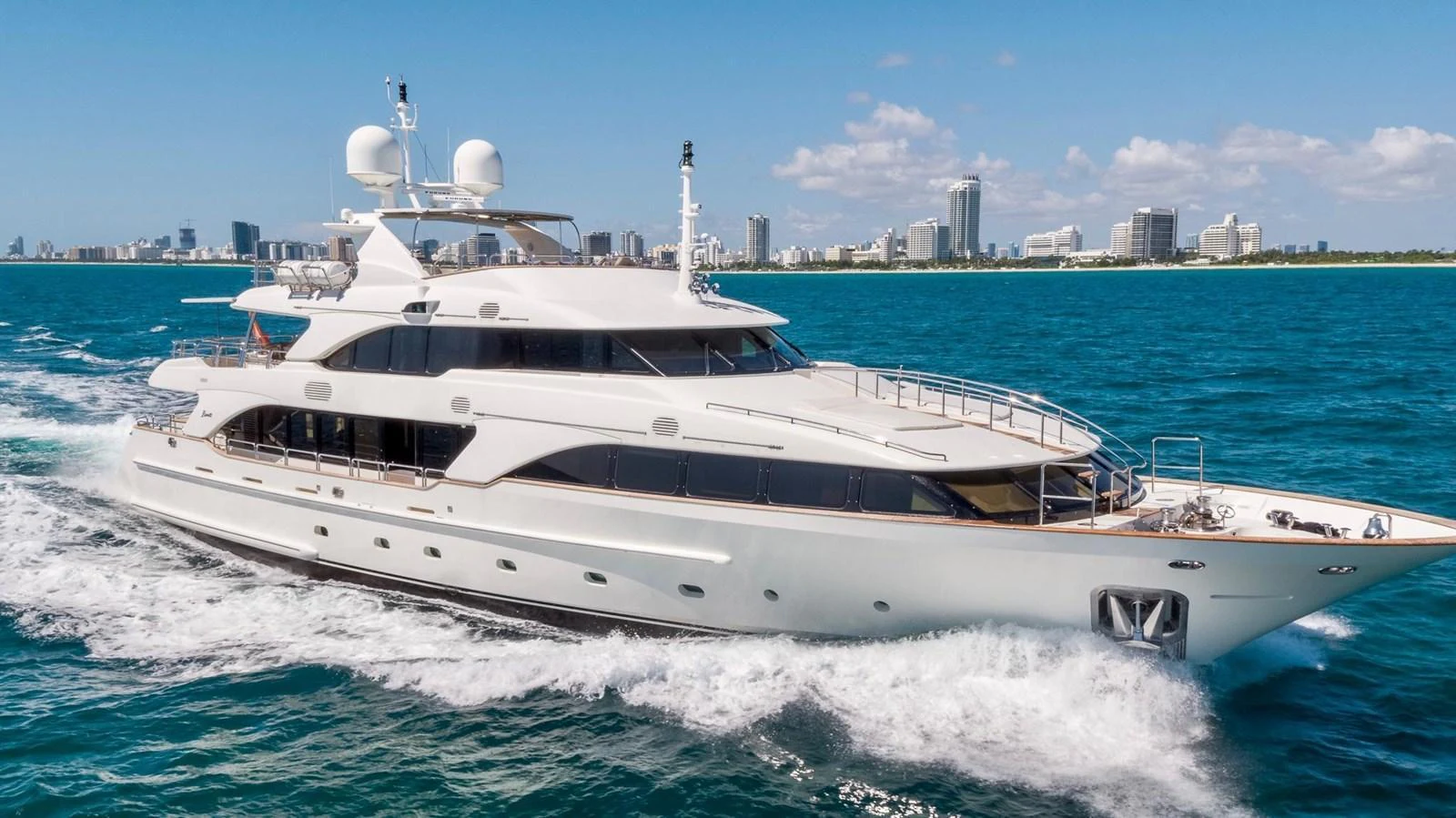 A sleek and luxurious yacht cruising through clear blue waters with a city skyline in the background. aboard BENETTI CLASSIC Yacht for Sale