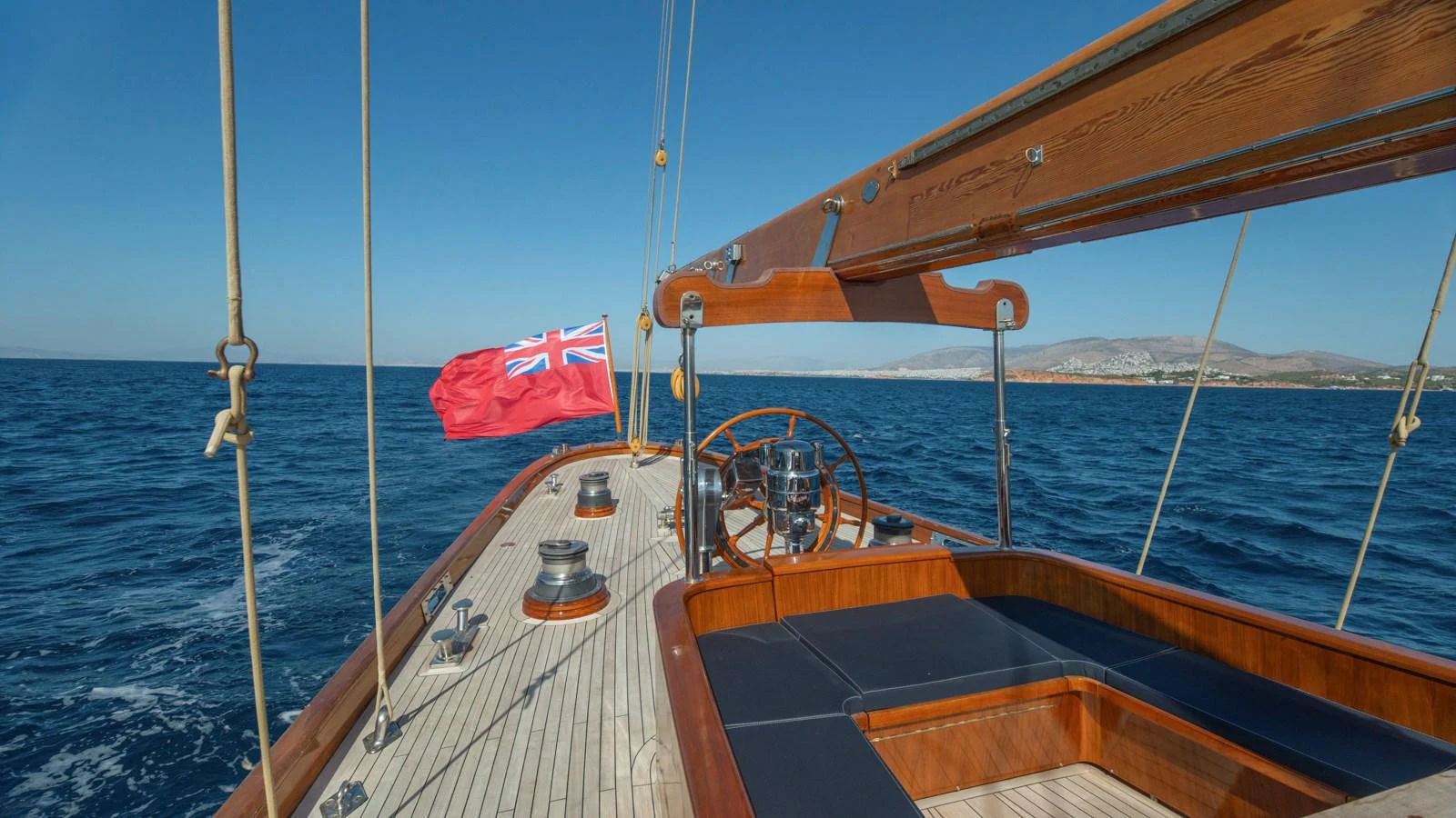 The yacht's deck features elegant wooden finishes and a classic steering wheel, with a vibrant flag waving in the breeze. aboard CANDIDA Yacht for Sale