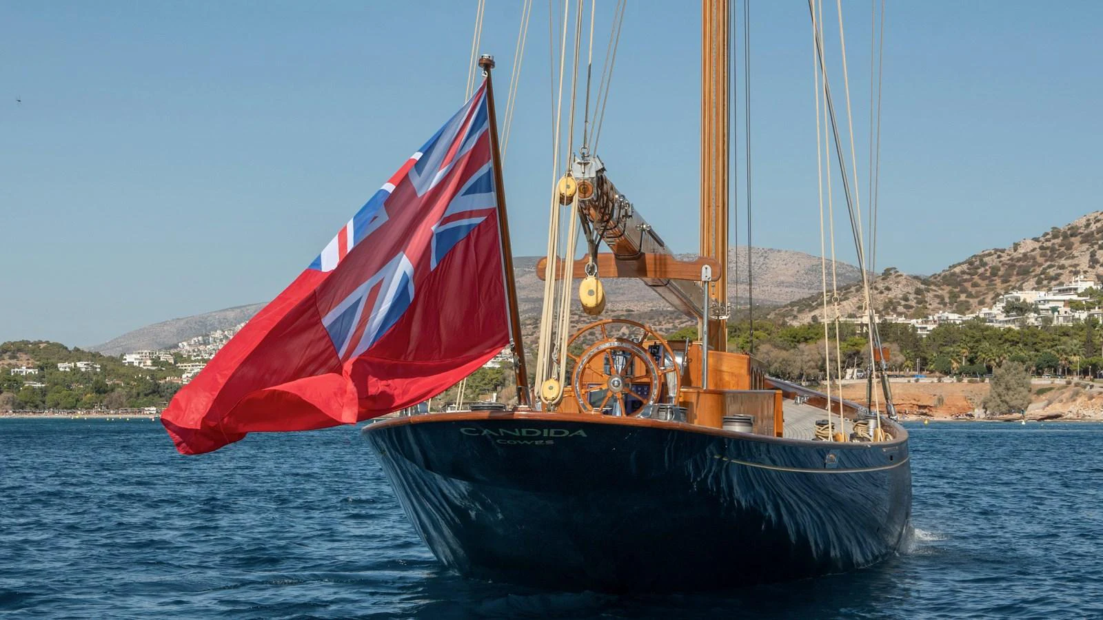 A classic yacht with a prominent red ensign flag sails on calm waters near a coastal landscape. aboard CANDIDA Yacht for Sale