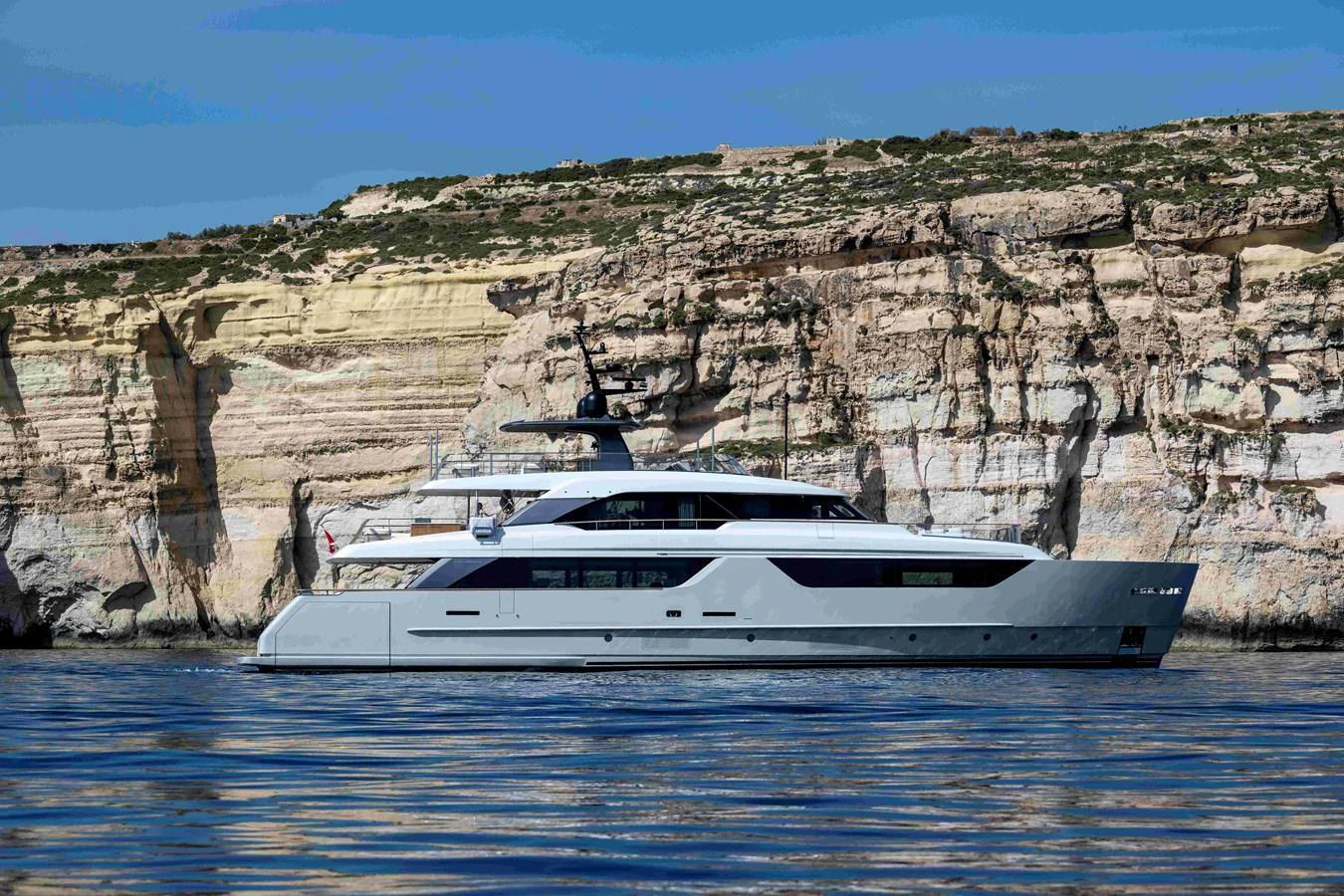 A sleek and modern yacht cruising along a rocky coastline under a clear blue sky. aboard UNIQUE S Yacht for Sale