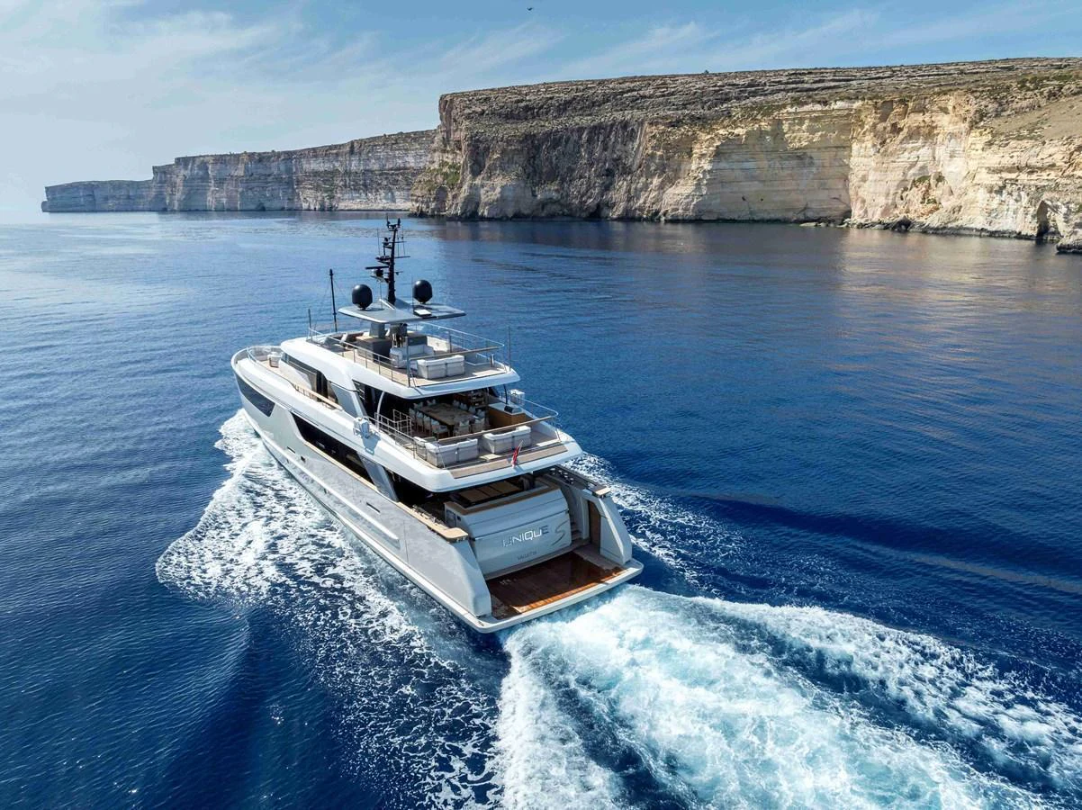 A sleek yacht cruises through pristine blue waters with stunning cliffs in the background. aboard UNIQUE S Yacht for Sale