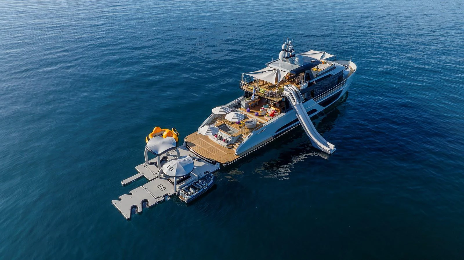 A luxurious yacht anchored on calm waters, featuring a slide, lounging areas, and a tender docked at the stern. aboard H2O - SPRITZ 116 Yacht for Sale