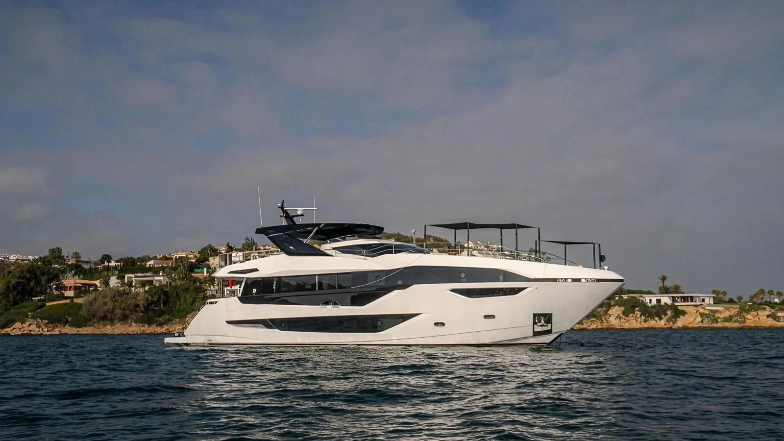 A sleek, modern yacht cruises along the coastline under a partly cloudy sky. aboard CONTESSA Yacht for Sale