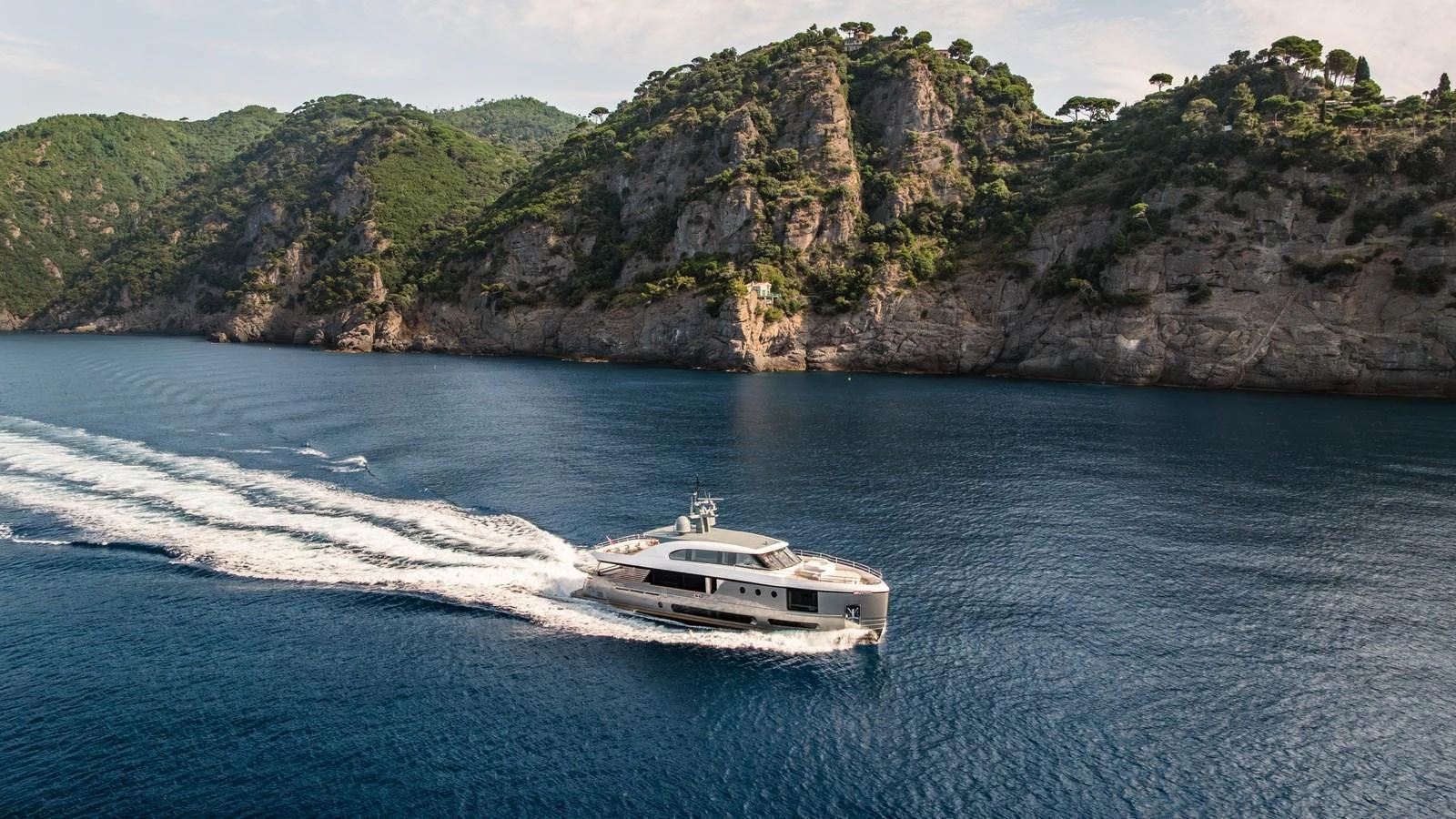 A sleek yacht cruises through serene blue waters with a stunning rocky coastline in the background. aboard ARIA Yacht for Sale