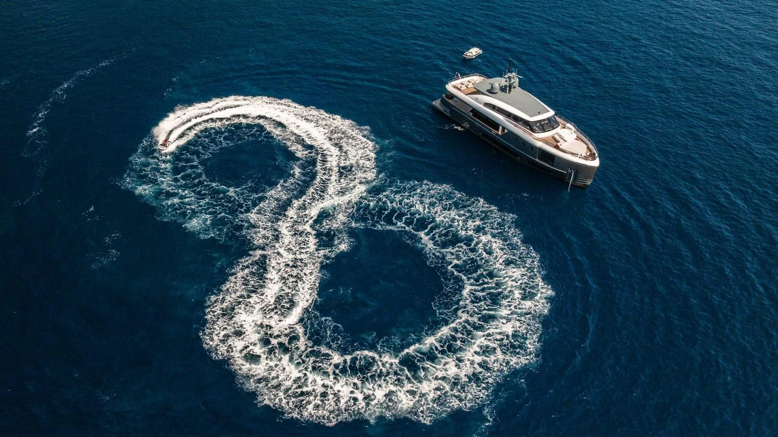 A sleek yacht cruises on the open sea, accompanied by a smaller watercraft creating dynamic patterns in the water. aboard ARIA Yacht for Sale