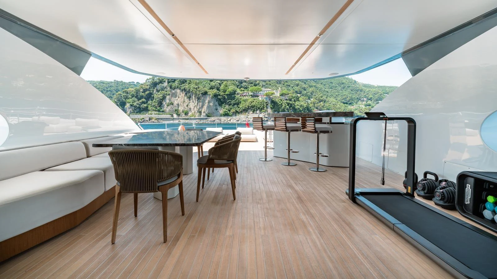 The aft deck features a spacious seating area, dining table, and exercise equipment with a scenic view. aboard ARIA Yacht for Sale
