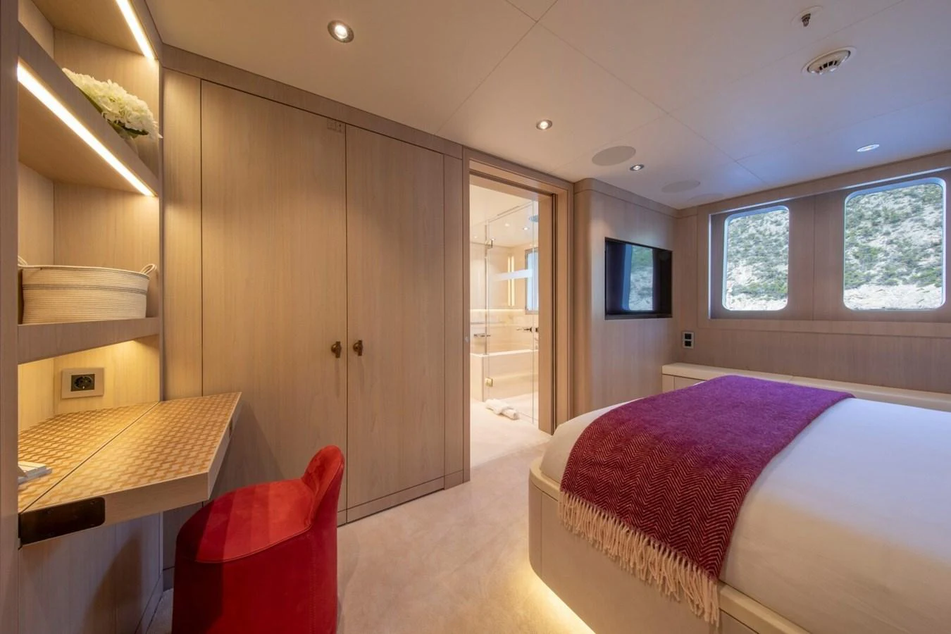 A luxurious stateroom featuring a cozy bed with a red throw, a desk area, and ample natural light. aboard PROMISE Yacht for Sale