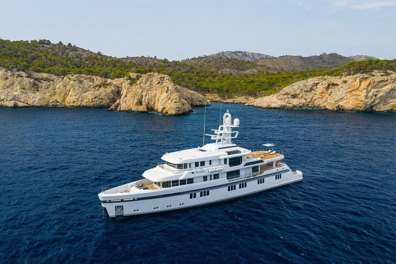 A sleek white yacht cruises along a serene coastline with rocky cliffs and lush greenery in the background. aboard PROMISE Yacht for Sale