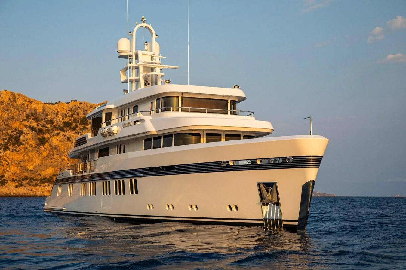 A luxurious yacht is anchored in serene waters against a backdrop of rocky cliffs at sunset. aboard PROMISE Yacht for Sale