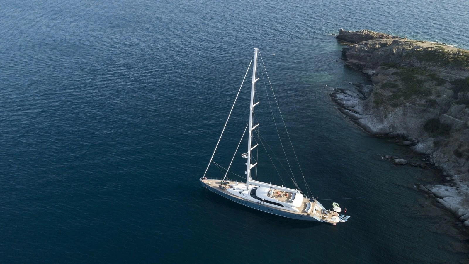 A sleek sailing yacht glides gracefully near a rocky coastline, showcasing its elegant design and spacious deck. aboard ALL ABOUT U Yacht for Sale