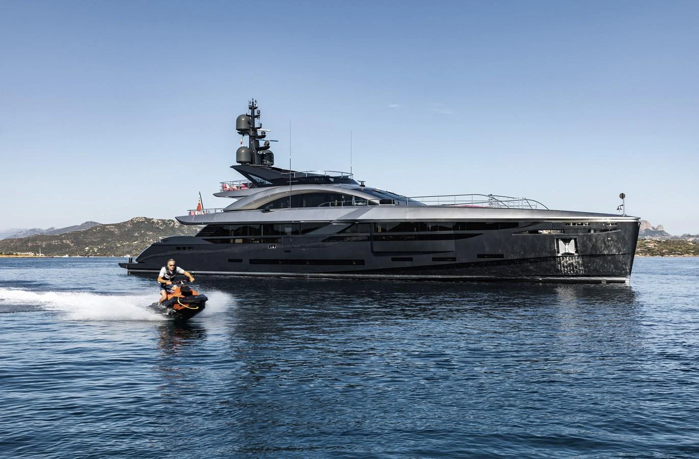 A sleek, modern yacht glides through calm waters under a clear sky, with a person riding a jet ski nearby. aboard FLYING DAGGER III Yacht for Sale