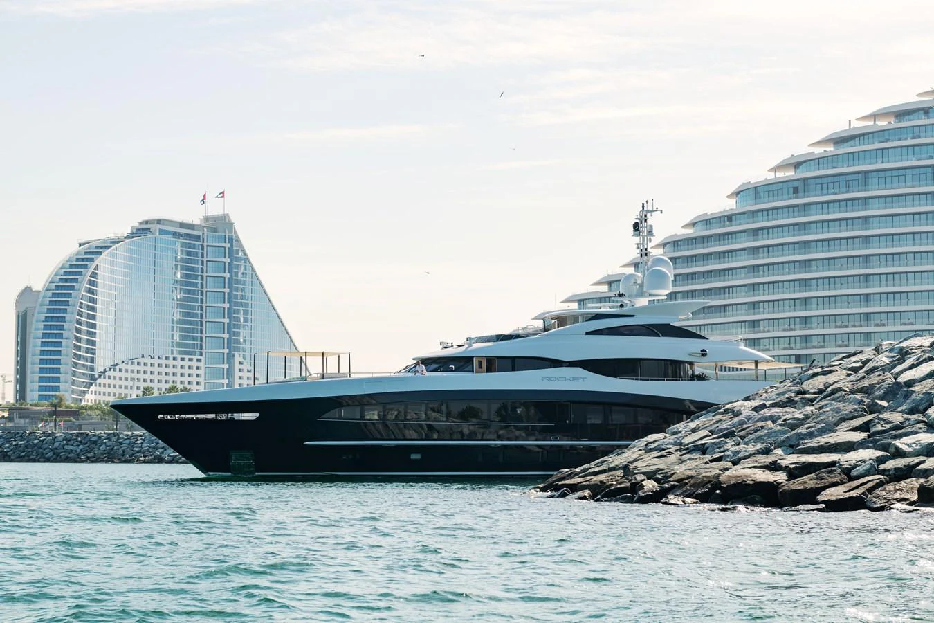 A sleek yacht is docked near a modern waterfront with luxurious buildings in the background. aboard ROCKET Yacht for Sale