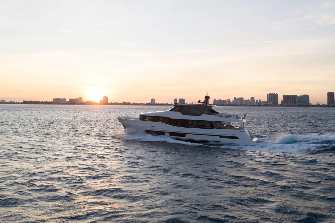 A sleek yacht cruises through calm waters at sunset with a city skyline in the background. aboard OCEAN ALEXANDER 28L Yacht for Sale