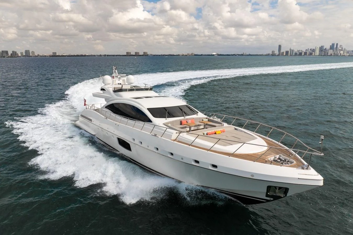 A sleek, modern yacht cruises swiftly through the water with a city skyline in the background. aboard SAYONARA Yacht for Sale
