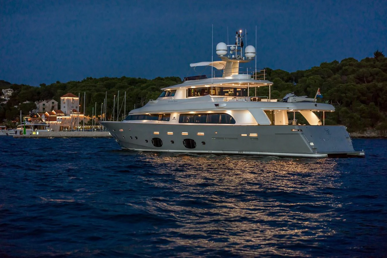 A luxurious yacht is anchored near a scenic coastline, illuminated beautifully against the evening sky. aboard SEVENTH SENSE Yacht for Sale