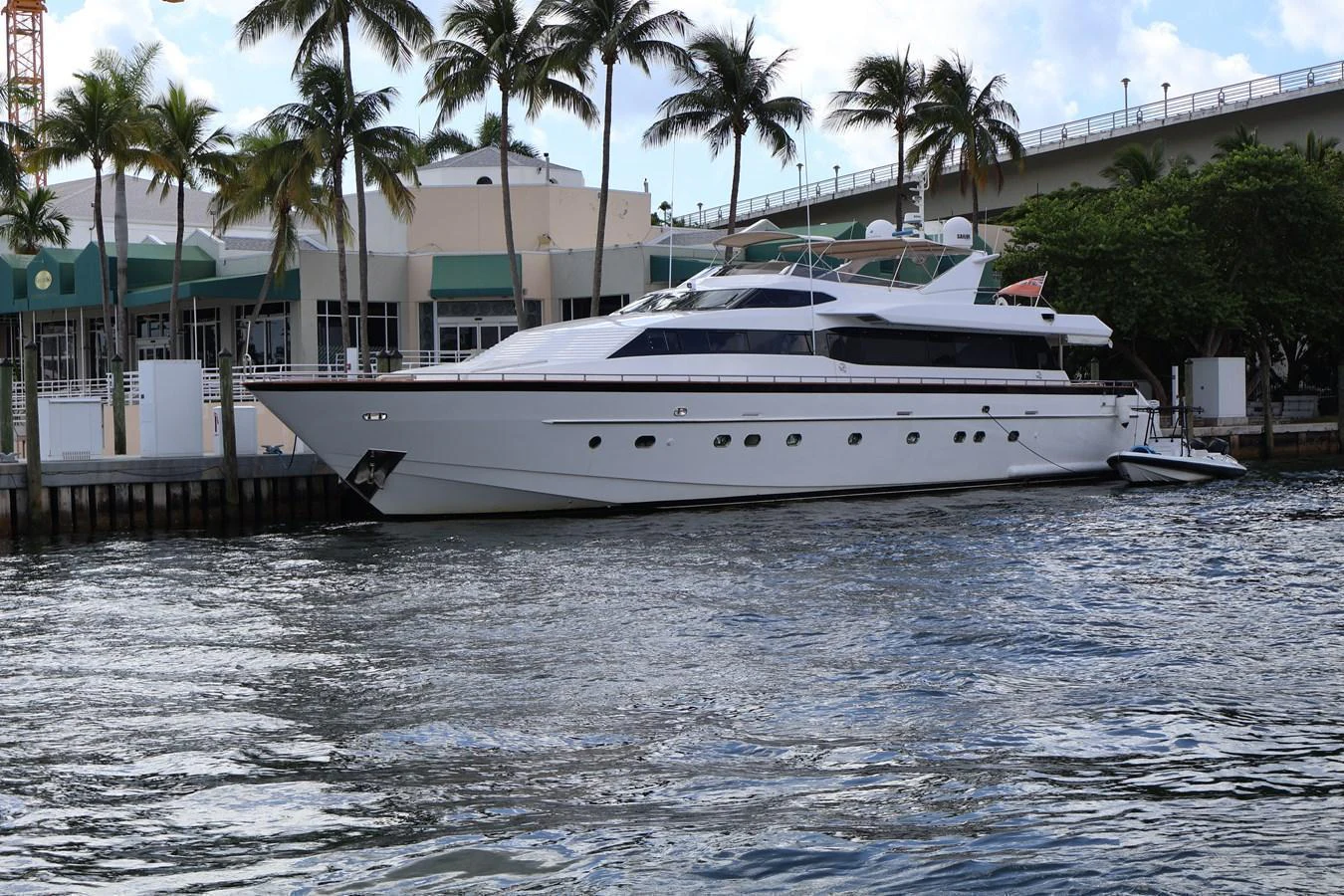 A sleek white yacht is docked by the waterfront, surrounded by palm trees and buildings. aboard Q Yacht for Sale