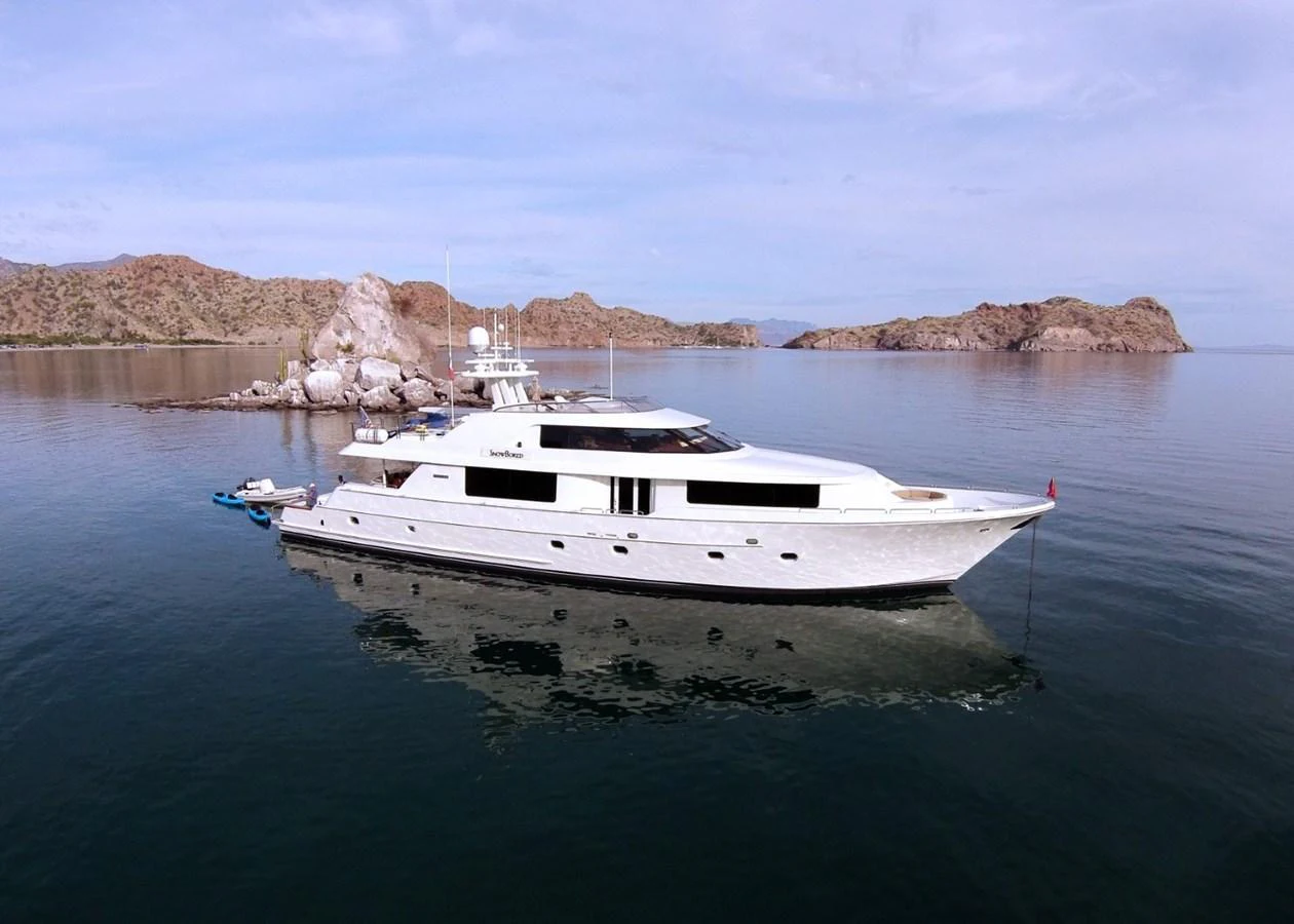 A sleek yacht is anchored in calm waters near a rocky coastline. aboard SNOWBORED Yacht for Sale