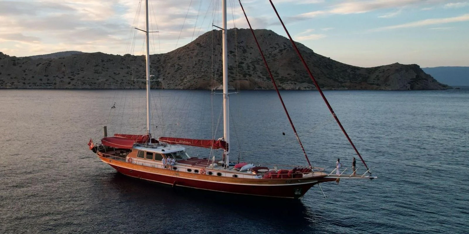 A classic sailing yacht gracefully anchored in a serene bay with scenic hills in the background. aboard AKHENATON Yacht for Sale