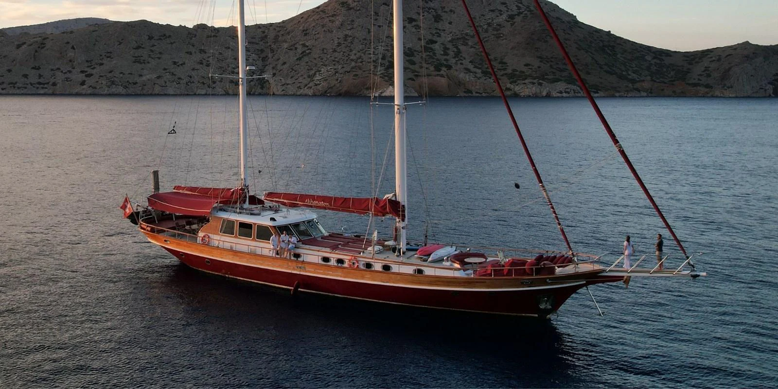 A classic sailing yacht with elegant lines is anchored in a serene bay at sunset. aboard AKHENATON Yacht for Sale