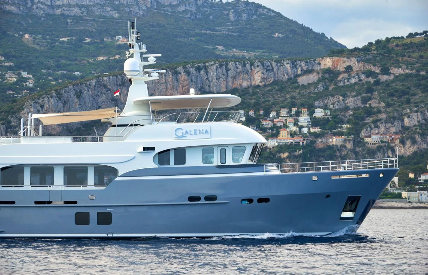 A sleek yacht cruises along a scenic coastline with a mountainous backdrop. aboard GALENA Yacht for Sale